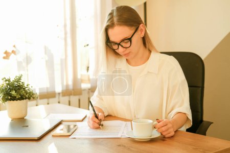 Téléchargez les photos : Image of smiling beautiful blond woman writing down notes while sitting at table in office. Lifestyle, people and business concept. - en image libre de droit