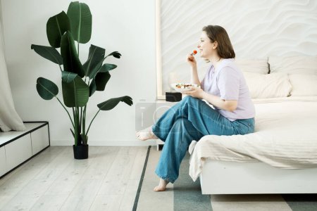 Téléchargez les photos : Pretty adult oversized woman sitting on bed and eating fresh vegetable salad - en image libre de droit
