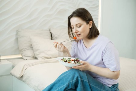 Foto de Mujer bastante adulta de gran tamaño sentada en la cama y comiendo ensalada de verduras frescas - Imagen libre de derechos