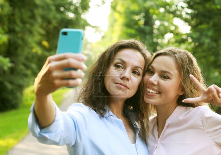 Photo for Mature mother and adult daughter are doing selfie by mobile phone in summer park - Royalty Free Image
