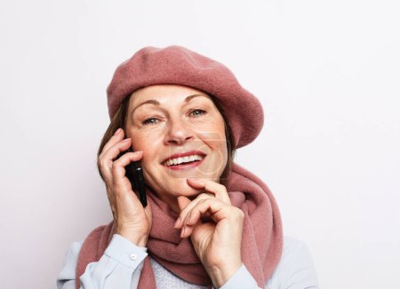 Téléchargez les photos : Femme âgée portant une écharpe rose et un chapeau avec smartphone sur fond gris blanc. - en image libre de droit