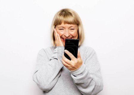 Photo for Pretty senior female in casual outfit using modern smartphone and laughing out loud while standing on white background. Old woman watching funny videos on internet. - Royalty Free Image