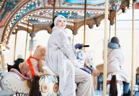 Téléchargez les photos : Charming woman in a faux fur coat and mittens rides a carousel and has fun - en image libre de droit