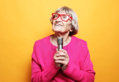 Photo for Emotion, lifestyle and old people concept: Happy old woman singing with microphone over yellow color background, having fun, expressing musical talent - Royalty Free Image