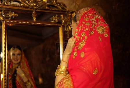Photo for A beautiful woman in a red sari, traditional Indian wedding dress, the bride looks in the mirror and admires her reflection. Woman covered with headscarf, indian culture. - Royalty Free Image