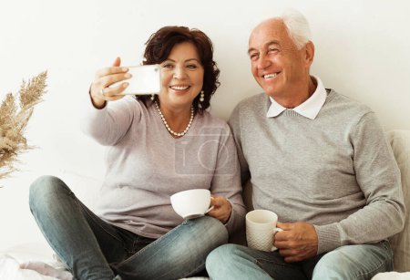 Foto de Caucasian senior man and woman woman on retirement. Happy couple sitting on bed and posing with smiles to phone camera at home. Old couple drinking coffee and taking selfie photos with smartphone. - Imagen libre de derechos