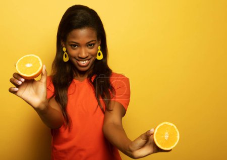 Téléchargez les photos : Lifestyle, food, diet and people concept: Optimist Young female model of afro appearance. Clean, even skin of the face. In the hands of citrus, orange. Photo on yellow background. - en image libre de droit