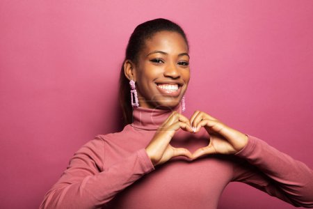 Foto de Young African American woman confesses in love, makes heart gesture, shows her true feelings, has happy expression, poses over pink background. Relationship concept. - Imagen libre de derechos