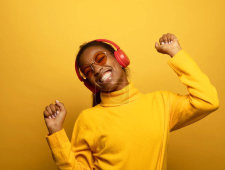 Foto de People, music, emotions concept. Young afro american female with dances in rhythm of melody, closes eyes listens loud song in headphones over yellow background. - Imagen libre de derechos