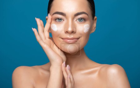 Téléchargez les photos : Beautiful young woman applying cosmetic cream treatment on her face isolated on blue color background - en image libre de droit