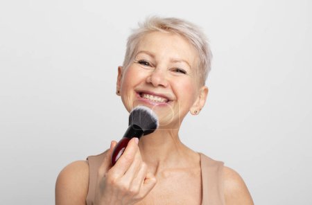 Photo for Beauty portrait mature woman applying blush with a makeup brush over grey background. - Royalty Free Image