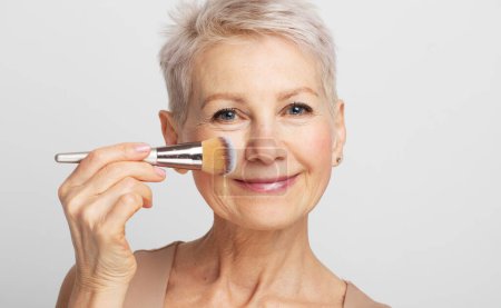 Téléchargez les photos : Portrait of beautiful smiling elderly woman doing makeup, applying blush to her cheek and looking at camera, light background with free space - en image libre de droit