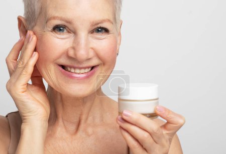 Foto de Smiling 60s middle aged mature woman putting tightening facial cream on face looking at camera. Anti age healthy dry skin care beauty therapy concept, skincare rejuvenation treatment against wrinkles. - Imagen libre de derechos