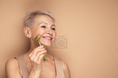 Photo for Gorgeous woman of 60s massaging face skin using natural facial stone trendy jade green quartz roller device for detox of perfect skin. Spa technique skincare ads. - Royalty Free Image