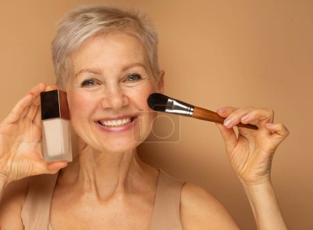 Photo for Pretty elderly lady holds makeup brush and foundation in her hands, prepares to do makeup. Close up portrait over beige background. - Royalty Free Image