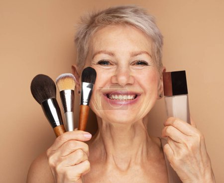 Téléchargez les photos : Charming elderly woman holds makeup brushes and foundation in her hands. Beauty at any age. - en image libre de droit