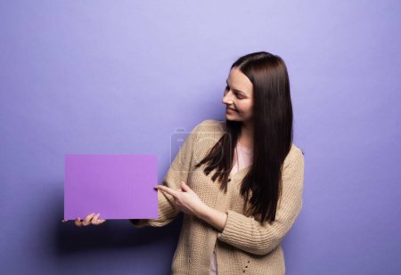 Photo for Beautiful young woman with long hair holding purple blank advertising board, smiling female demonstrating empty banner with copy space for your text or design, standing on lilac background in studio - Royalty Free Image