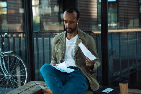 Téléchargez les photos : A young African American man sits at a bus stop and reads documents. Lifextyle concept. - en image libre de droit