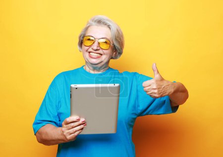 Foto de Education, old people and tehnology concept: Elderly woman holding an ipad and showing thumbs up sign over yellow background - Imagen libre de derechos