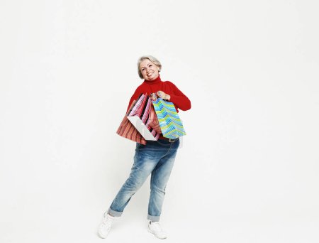 Foto de Full length photo of positive cheerful grey white hair old woman enjoy leisure time shopping hold bags wear red sweater and jeans, posing over white background. - Imagen libre de derechos