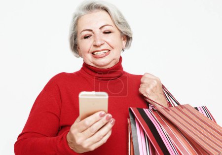 Téléchargez les photos : A charming old woman with white hair holds shopping bags in her hands and checks discounts on her smartphone. Modern technologies for shopping. - en image libre de droit