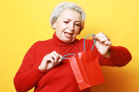 Téléchargez les photos : A cheerful and joyful old woman with gray hair holds a red bag in her hands and looks into it. The joy of a gift. - en image libre de droit