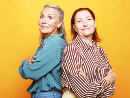 Foto de Two pretty elderly women friends with crossed arms on yellow background. Lifestyle concept. - Imagen libre de derechos
