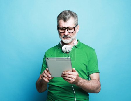 Photo for Lifestyle, tehnology and people concept: Happy senior bearded man wearing eyeglasses and headphones using digital tablet. Studio shoot over blue color background. - Royalty Free Image