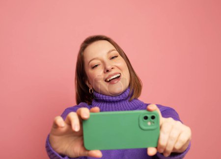 Téléchargez les photos : Cheerful smiling funny woman 40s wearing casual sweater posing doing selfie shot on mobile phone on pastel pink colour background studio portrait - en image libre de droit