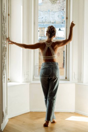 Photo for Young blond woman in jeans and a lace top is standing in the living room, near big window, back view - Royalty Free Image