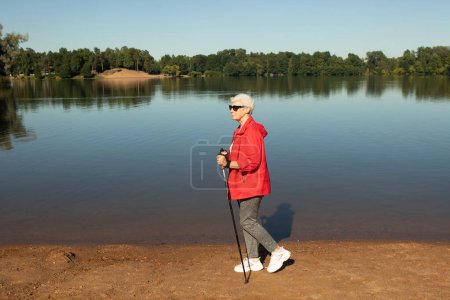 Foto de Mujer de pelo gris caminando con palos de seguimiento en la playa cerca del lago, concepto de estilo de vida. - Imagen libre de derechos