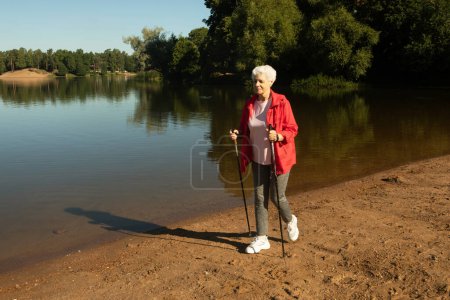 Téléchargez les photos : Grey-haired woman walking with tracking sticks on the beach near lake, lifestyle concept. - en image libre de droit