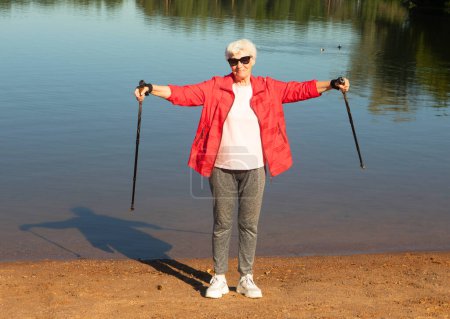 Téléchargez les photos : Grey-haired woman in sunglasses walking with tracking sticks on the beach near lake. Sport, healthy lifestyle and happy old age concept. - en image libre de droit