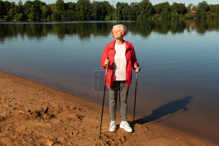 Téléchargez les photos : Summertime, active lifestyle, leisure and hobby concept. Outdoor shot of healthy energetic elderly female in red jacket and headphones walking on the beach sunny day using Nordic poles. - en image libre de droit