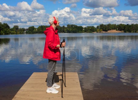 Téléchargez les photos : An elderly woman with a short haircut dressed in red sportswear holds walking sticks in her hands and listens to music on headphones, standing on the shore of a lake - en image libre de droit
