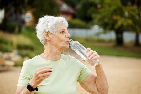 Photo for Senior woman with short grey hair drinking water after exercising, summer time, portrait in the park - Royalty Free Image
