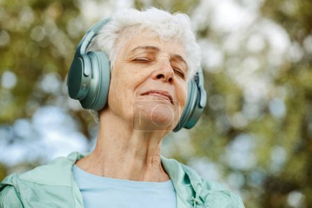 Photo for Elderly woman in headphones listens to music in the park, concepts about elderly, seniority and wellness aging. Summer time. - Royalty Free Image