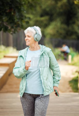 Foto de An elderly woman with a short haircut dressed in sportswear is jogging in the park while listening to music on headphones. Lifestyle and old people concept. - Imagen libre de derechos