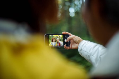 Téléchargez les photos : Multiracial elderly women having fun during trekking day in to the wood and make selfie with smartphone. View from the back. - en image libre de droit