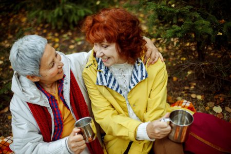 Foto de Two elderly women are sitting on a plaid blanket in the forest, drinking coffee, talking, having fun. Lifestyle concept. - Imagen libre de derechos