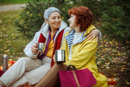 Téléchargez les photos : Two elderly women are sitting on a plaid blanket in the forest, drinking coffee, talking, having fun. Lifestyle concept. - en image libre de droit