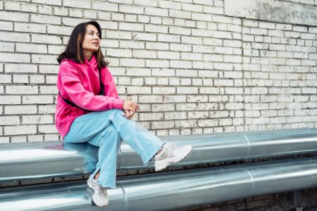 Téléchargez les photos : Fashion Asian young woman in a pink sweatshirt and jeans sits against a brick wall background, has fun. - en image libre de droit