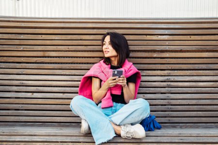 Foto de A fashionable young woman sits on a bench and holds a smartphone. Asian beauty and fashion. Summer time. - Imagen libre de derechos