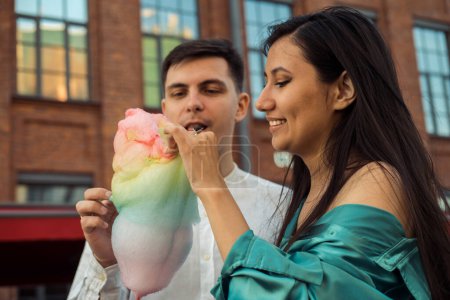 Téléchargez les photos : A cheerful couple in love walks in the city and eats cotton candy. Young Asian woman with long hair and European man. Summer day. - en image libre de droit