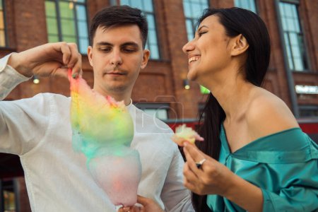 Foto de A cheerful couple in love walks in the city and eats cotton candy. Young Asian woman with long hair and European man. Summer day. - Imagen libre de derechos