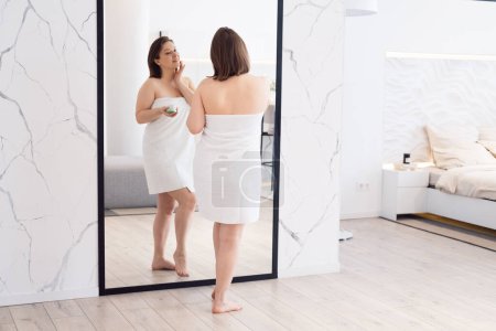 Téléchargez les photos : Adult chubby woman apply body cream look in mirror at home. Beauty and lifestyle concept. - en image libre de droit