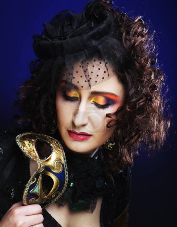 Foto de Beautiful curly woman with bright makeup and hat with veil holding venetian mask over blue color background - Imagen libre de derechos