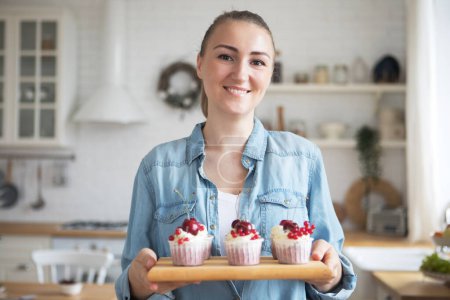 Photo for Work in confectionery workshop. Handmade cupcakes. Smiling pretty young woman pastry chef, demonstrating tasty fresh cupcales with berries to camera. - Royalty Free Image