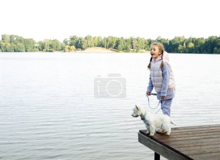 Photo for A seven-year-old girl with a small white dog stands on a bridge near the lake and looks at the water. Friendship with pets. Happy childhood. - Royalty Free Image