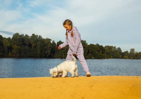 Téléchargez les photos : A seven-year-old girl with pigtails plays with a west highland white terrier on the beach near the lake. Lifextyle concept. - en image libre de droit